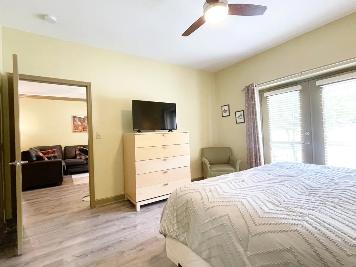 A serene master bedroom scene featuring a king-sized bed with a plush mattress, a dresser, a TV, a small seating area, and doors that lead outside. The room is bathed in soft light, enhancing its comfortable and peaceful ambiance.