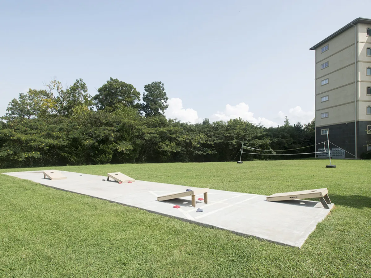 The image features an open grassy area with cornhole boards set up for play. The condo building is visible in the background, and the scene conveys a sense of playful outdoor activity and community gathering space.