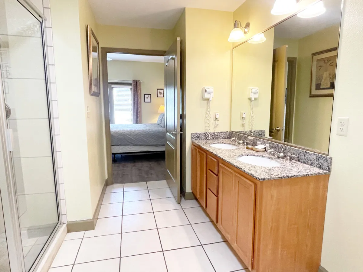 A calm and functional bathroom featuring a double vanity with granite countertop, a spacious shower enclosure, and a doorway leading toward the bedroom, evoking a sense of comfort and luxury.
