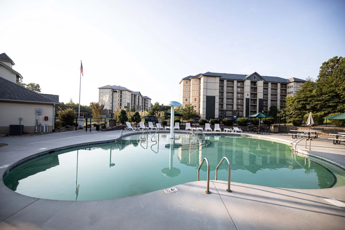 The image displays a sparkling swimming pool surrounded by lounge chairs, with the condo buildings in the background and an American flag waving nearby. The mood is lively and social, suggesting summer fun and family gatherings by the water.