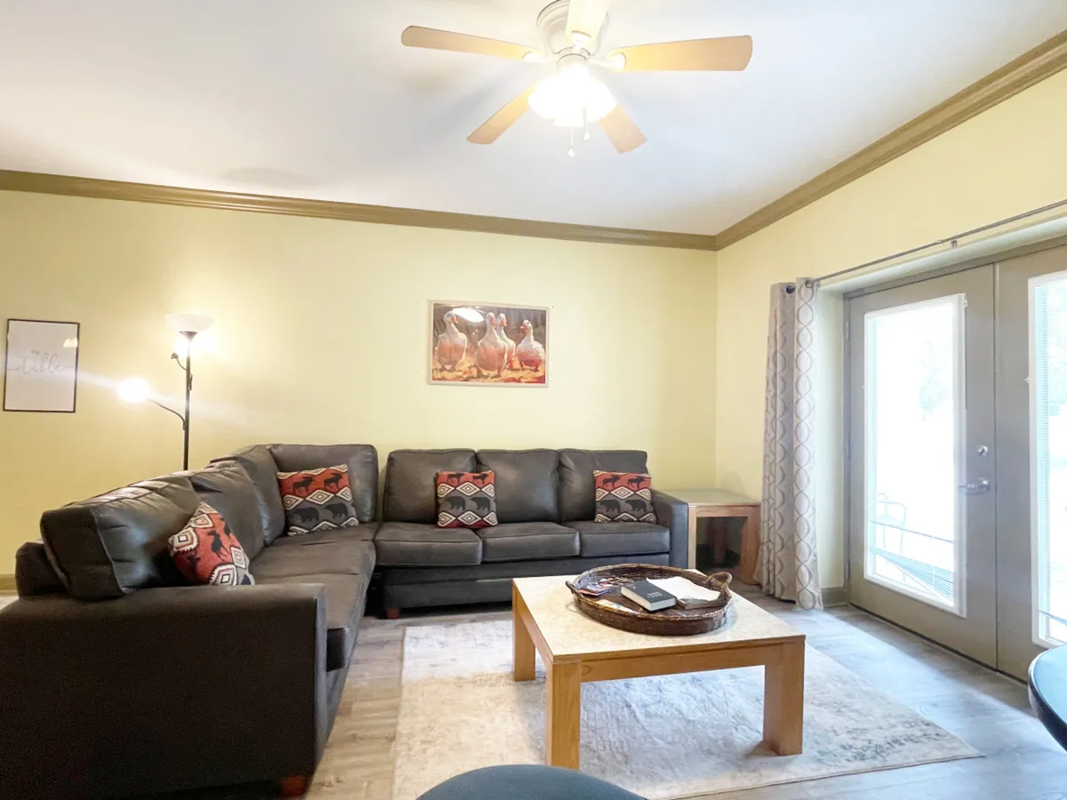 A cozy and inviting living room with a sectional sofa, coffee table, wall artwork, and a warm light fixture, suggesting a space ideal for relaxing, intimate conversations, or unwinding after an eventful day.