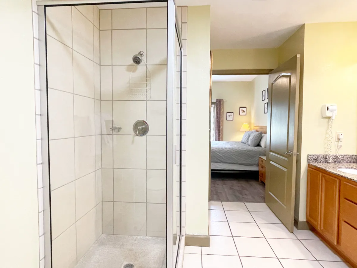 A view of a modern bathroom focusing on a glass shower enclosure, granite countertop, and an open doorway leading to a cozy bedroom. The scene invites a sense of comfort, with soft lighting and soothing decor.