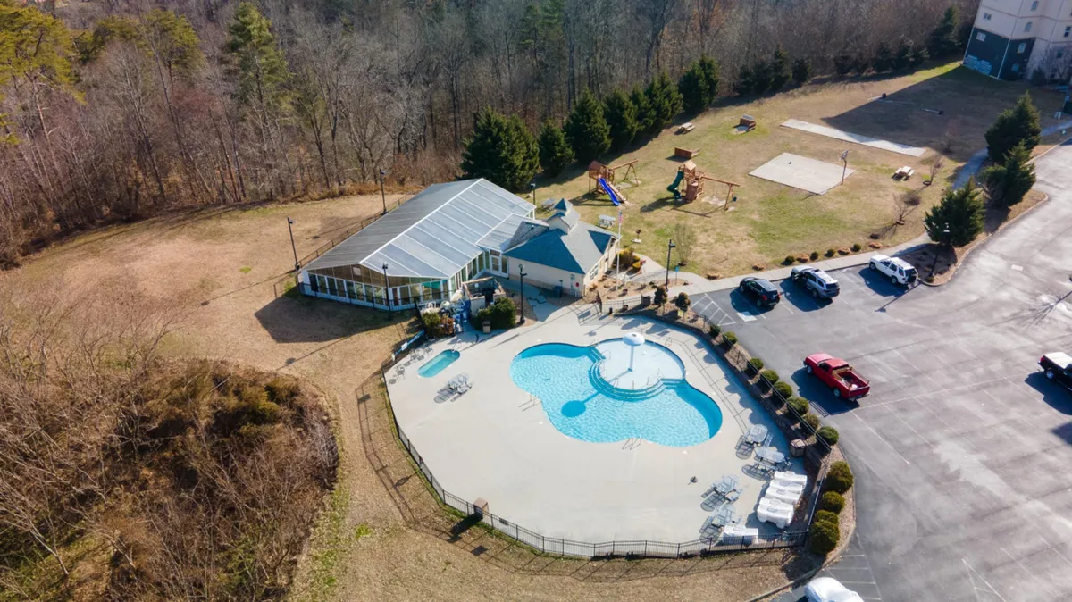 This exterior image shows a large swimming pool surrounded by a recreational area and visible playground equipment, with a parking lot in the background. The poolside area is designed for families, with ample space for children to play and adults to relax. The presence of the playground highlights the family-friendly amenities.