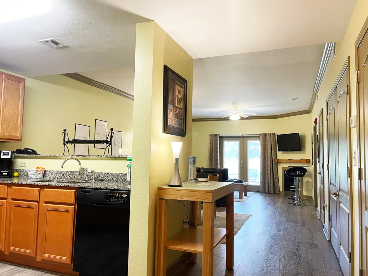 A wider shot encompassing the kitchen area, dining table, inviting living area, and a window with curtains. The space is set up for cooking, dining, and socializing, offering a sense of togetherness and communal activity.