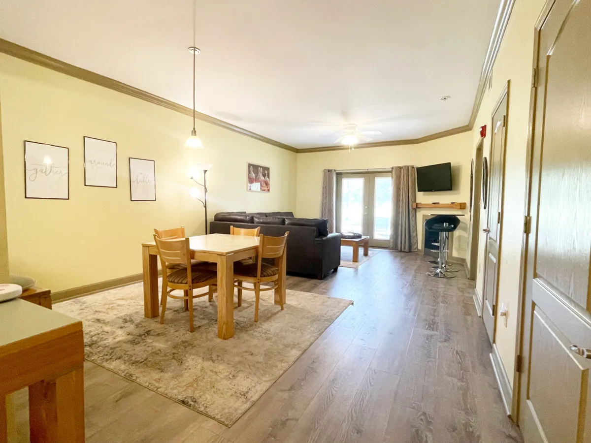 The image showcases a cozy and welcoming living area with a plush sofa and a large flat-screen TV. Large windows fill the space with natural light, while the dining table is positioned nearby, indicating an open layout that encourages togetherness and easy flow between the living, dining, and kitchen spaces.